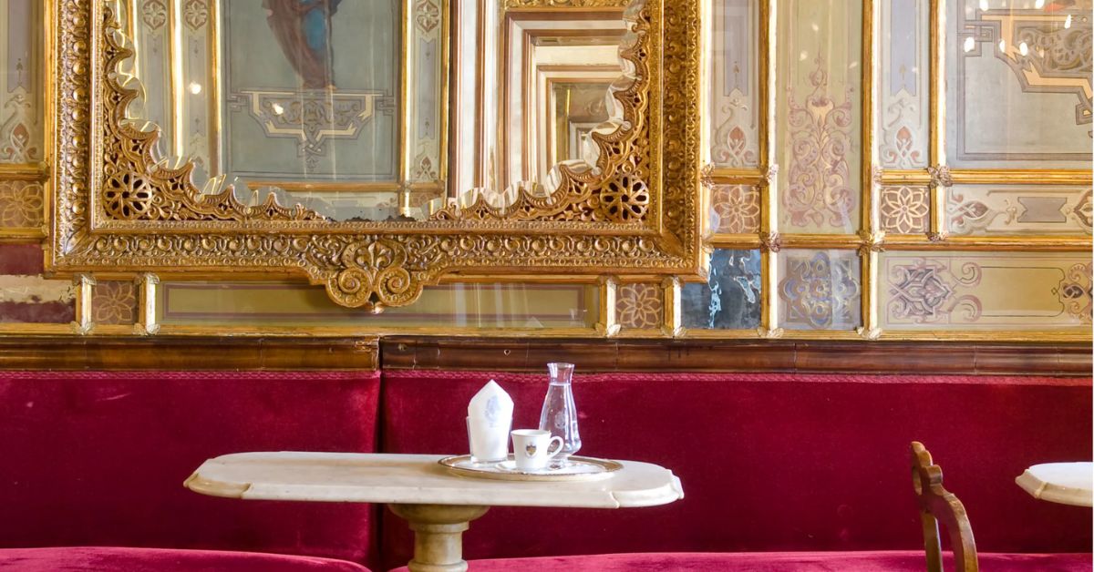a beautiful white marble café table in front of a red velvet banquette with a 300 year old gold antique mirror wall behind it. This is Caffè Florian in Venice, Italy. The oldest cafe in the world.