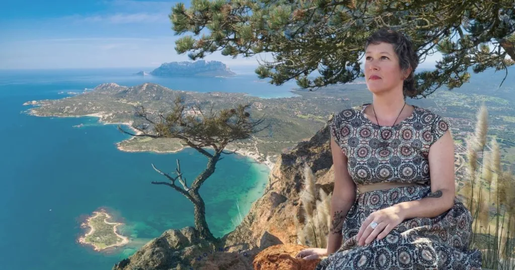 Author Jenn Campus sitting at the top of a cliff looking out over the turquoise waters of Sardinia. The island of Sardinia is visible in the background and Jenn is surrounded by native Sardinian pine trees and grasses. Jenn is wearing a crystal necklace and a grey and navy blue dress with Moroccan pattern tied with a brown belt. Jenn is seated gazing out to the horizon with knees bent and pulled toward her with one hand in her lap and her other hand is resting on a orange clay-colored rock.