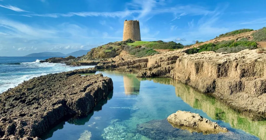 the clear water of the Mediterranean Sea on the coast of the Island of Sardinia with the ruins of an ancient stone tower reflected in the crystal clear water. There is a clear blue sky with small clouds and the flora and fauna of Sardinia