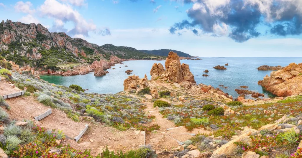 the coast of Sardinia with clear water and blue sky and clouds. The hills of the island are covered in local flora and fauna and rocks.