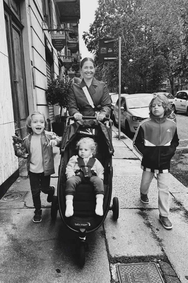 A smiling woman pushing a stroller in Turin Italy with a little blonde boy in the stroller and two older blonde boys walking on either side of her - Barrett and the Boys